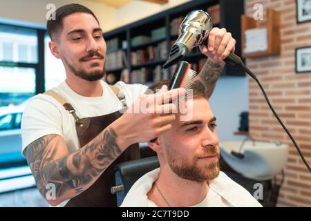 Coiffeur jeune peignant et sécher les cheveux du client mâle dans un barbershop Banque D'Images