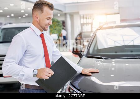 portrait de jeune consultant de voitures salle d'exposition, homme caucasien se tient près de l'auto noire, tenant des documents de voiture dans les mains Banque D'Images