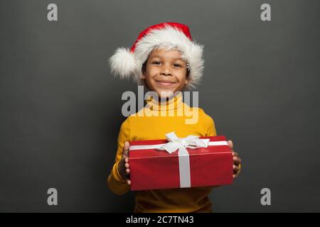 Père Noël enfant noir tenant une boîte de Noël rouge avec ruban blanc soyeux sur fond gris Banque D'Images