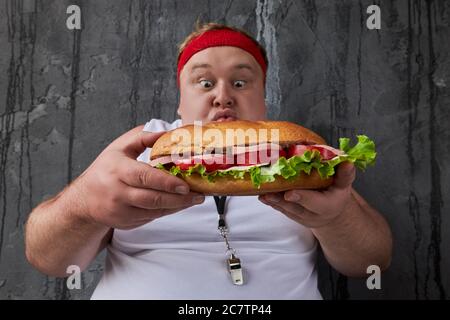 un jeune homme de caucase en surpoids empiète sur un grand sandwich à haute teneur en calories, l'homme affamé veut manger, il ne s'occupe pas de gain de poids Banque D'Images