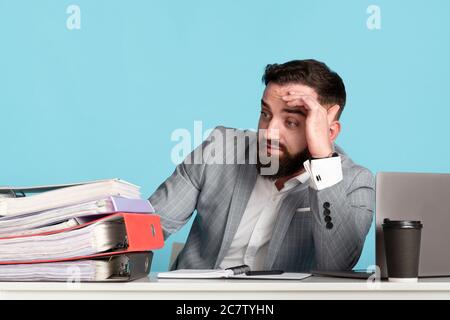 Un homme d'affaires millénaire épuisé a été submergé de documents administratifs à son bureau avec un ordinateur portable sur fond bleu Banque D'Images