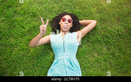 Belle fille noire couchée sur l'herbe verte, montrant le geste de paix et en collant sa langue. Vue de dessus Banque D'Images