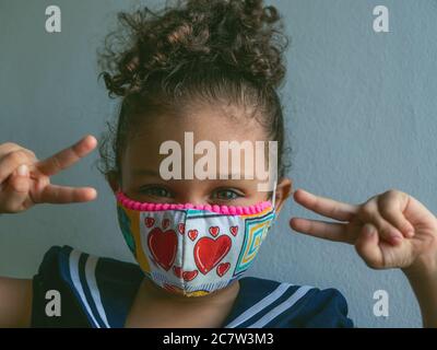 Une petite fille avec un masque de coeur regarde l'appareil photo avec ses doigts avec le signe de la paix Banque D'Images