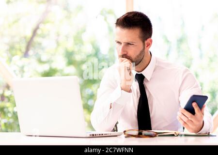 Photo d'un homme d'affaires élégant, assis derrière son ordinateur portable au bureau. Homme professionnel tenant un téléphone portable dans la main. Banque D'Images