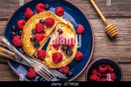 Tranches de crêpes maison sucrées avec framboises et bleuets sur une assiette bleue sur un bureau en bois sombre. Banque D'Images