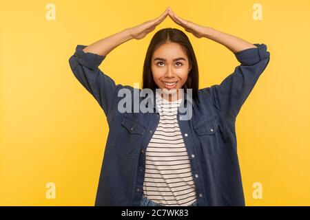 Je suis en sécurité ! Portrait de sympathique belle fille en chemise en denim tenant le geste de la main de toit sur la tête et souriant, se sentant sécurisé, rêvant de maison. Banque D'Images