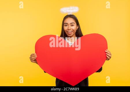 Portrait de la belle fille angélique avec Saint nimbus tenant grand coeur lu et souriant à l'appareil photo, exprimant l'amour, salutation le jour de la Saint-Valentin. Intérieur Banque D'Images