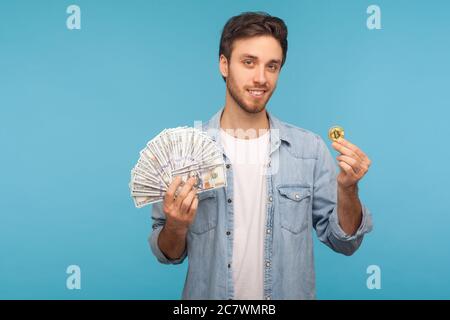 Crypto-monnaie, investissement électronique. Un homme charmant et heureux dans une chemise en denim ouvrier montrant des billets de banque et des bitcoins en dollars, regardant l'appareil photo avec le mottier en dents de main Banque D'Images