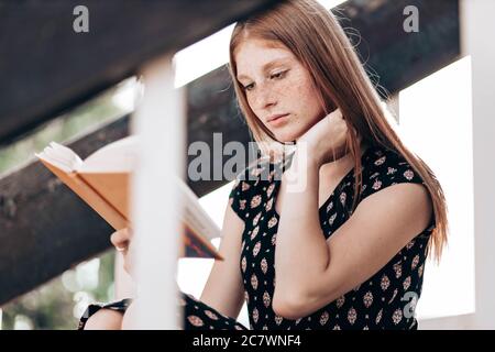 Jeune fille, gingembre, adolescente, assise sur les escaliers devant son école, lisant un livre. Banque D'Images