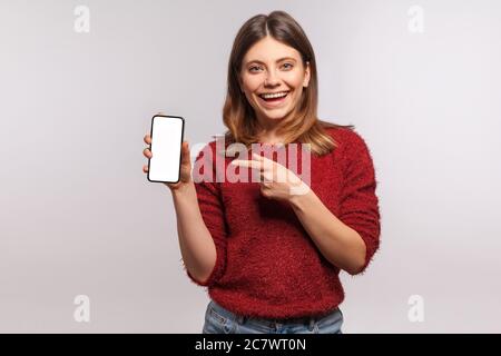 Regardez la publicité mobile ! Bonne femme souriante brune dans un pull tirateux pointant téléphone cellulaire avec écran vide, maquette vide pour le texte commercial Banque D'Images