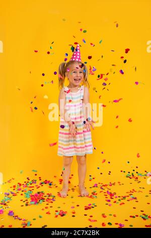 Joyeux anniversaire sourire enfant fille dans une tasse rose entouré de confetti volant sur fond jaune coloré . Célébration, enfance, émotions Banque D'Images