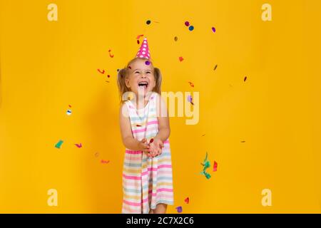 Joyeux anniversaire sourire enfant fille dans une tasse rose entouré de confetti volant sur fond jaune coloré . Célébration, enfance, émotions Banque D'Images