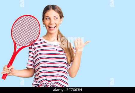 Belle femme caucasienne avec cheveux blonds jouant au tennis tenant raquette pointant le pouce vers le côté souriant heureux avec la bouche ouverte Banque D'Images