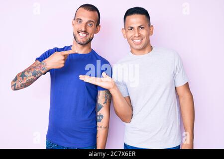 Jeune gay couple de deux hommes portant des vêtements décontractés stupéfaits et souriant à l'appareil photo tout en présentant avec la main et pointant avec le doigt. Banque D'Images