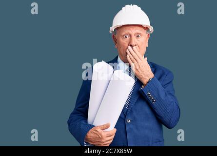 Homme senior beau gris-cheveux portant un casque d'architecte tenant des bleus couvrant la bouche avec la main, choqué et peur d'erreur. Surpris expre Banque D'Images