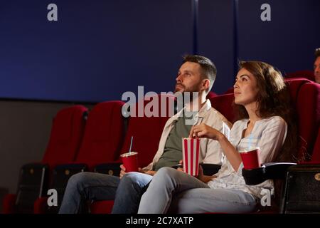 Portrait d'un jeune couple moderne en cinéma, regardant un film et appréciant le pop-corn tout en étant assis sur des chaises en velours rouge dans une pièce sombre, concentrez-vous sur une femme souriante Banque D'Images