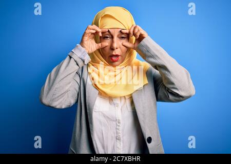Femme d'affaires brunette d'âge moyen portant le hijab traditionnel musulman sur fond bleu essayant d'ouvrir les yeux avec les doigts, endormi et fatigué pour la mornine Banque D'Images