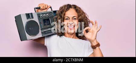 Moyen âge brunette hipster femme tenant la musique rétro boombox sur fond rose isolé faire signe ok avec les doigts, souriant sympathique gesting exc Banque D'Images