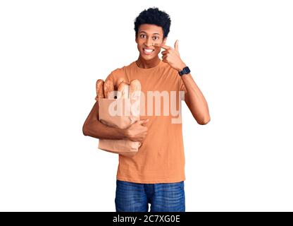 Jeune afro-américain tenant sac de papier avec pain pointant du doigt vers le visage et le nez, souriant gai. Concept de beauté Banque D'Images