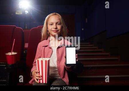 Portrait de la jeune fille blonde montrant un smartphone avec écran vierge et regardant l'appareil photo tout en tenant une tasse de pop-corn dans le cinéma, cop Banque D'Images