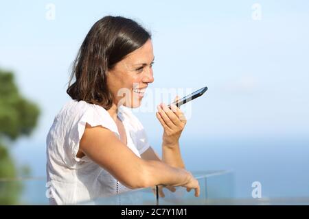 Bonne femme adulte utilisant la reconnaissance vocale sur un smartphone debout sur un balcon sur la plage Banque D'Images