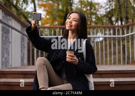 Jeune belle femme décontractée avec café à aller avec plaisir prendre selfie sur le téléphone portable dans le parc Banque D'Images