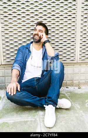 Homme à barbe gai dans une tenue décontractée assis sur le sol de la rue tout en se relaxant sur une pause-café Banque D'Images