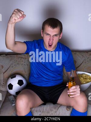 drôle jeune homme regardant le football à la télévision avec des jetons et de la bière et célébrant le but Banque D'Images