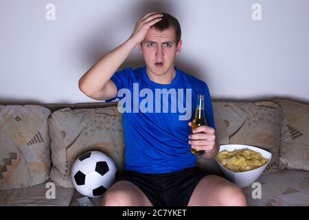 supporter choqué en uniforme bleu assis sur le canapé et regardant le football avec de la bière et des frites Banque D'Images