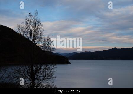 silhouette de montagnes et de branches d'arbres sur un large lac paisible. nuages blancs dans le ciel bleu au coucher du soleil. Dans le lac Lugu Yunnan Chine Banque D'Images