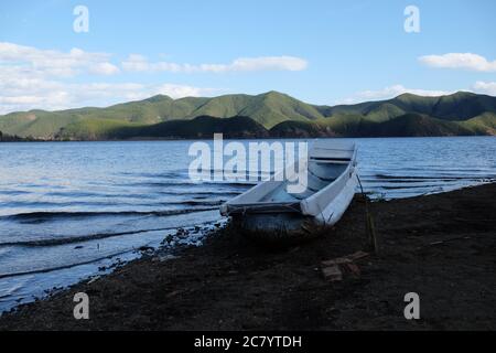 une rangée de bateaux blancs sur la plage de sol noir. Le lac bleu se déforme. Fond vert des montagnes. Ciel bleu ensoleillé. En Lugu lac Sichuan Chine. Banque D'Images