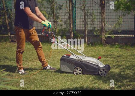 Homme dans une tenue décontractée et des gants est de couper l'herbe verte avec la tondeuse moderne sur son arrière-cour. Matériel et services de jardinage. Jour ensoleillé Banque D'Images