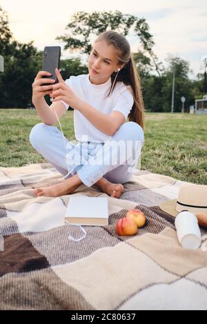 Belle adolescente dans des écouteurs prendre des photos sur téléphone portable lors d'un pique-nique dans le parc Banque D'Images