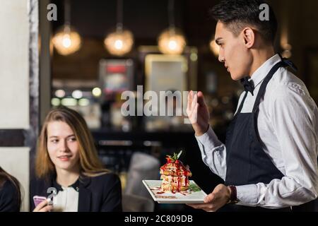 Un jeune serveur positif servant des clients du restaurant en terrasse à table.Focus sur l'homme Banque D'Images