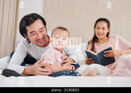 Jolie jeune femme avec livre entre les mains regardant son mari jouer avec un petit fils Banque D'Images