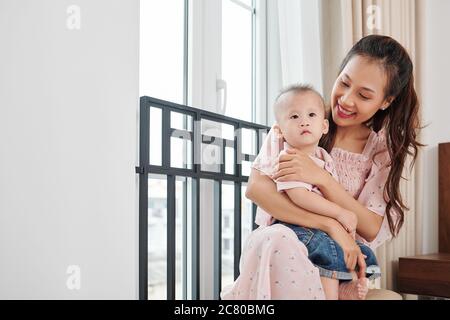 Belle jeune femme vietnamienne assise à côté de la fenêtre et tenant son petit fils Banque D'Images
