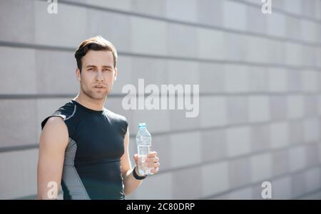 Formation, soif et eau. Un homme fatigué en vêtements de sport tient une bouteille à la main, sur fond de mur Banque D'Images