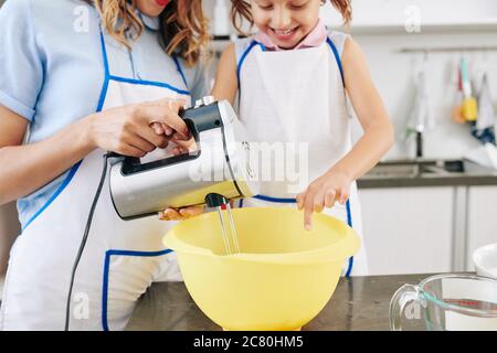 Bonne petite fille et sa mère mélangeant la pâte à crêpes dans un grand bol en plastique avec un mélangeur électronique Banque D'Images