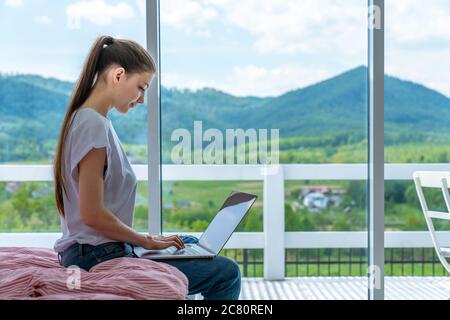 Vue rognée de la jeune femme Keyboard sur ordinateur portable avec écran vide de l'espace de copie tout en étant assis sur le lit à la maison, une femme brunette étudiante Banque D'Images