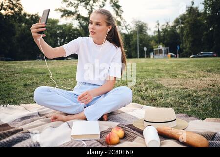 Une adolescente attirante dans des écouteurs prend des photos avec un téléphone portable tout en étant assise pour pique-niquer dans le parc de la ville Banque D'Images