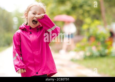 Petite fille hurlant debout seule et se perdre dans le parc à l'extérieur. Bébé pleurant portant un imperméable. Banque D'Images