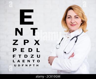 portrait d'une femme mature médecin ou infirmière posant sur un mur blanc avec un tableau de test oculaire Banque D'Images