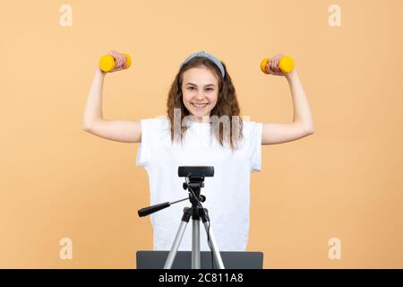 Happy girl blogger se tient devant la caméra avec des haltères et un ordinateur portable et enregistre la vidéo sur la caméra comme elle fait des exercices à la maison Banque D'Images