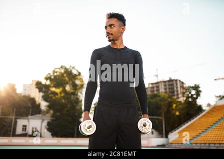 Jeune sportif afro-américain élégant avec haltères, qui regarde avec assurance l'entraînement au stade Banque D'Images