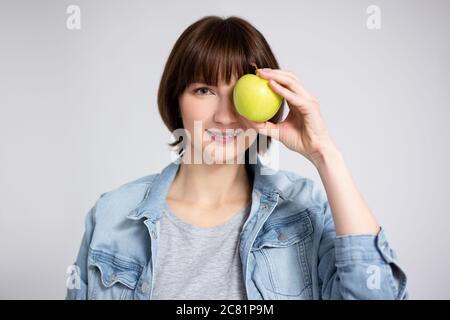 portrait d'une jeune femme ou d'une adolescente avec des bretelles sur les dents couvrant le visage avec une pomme verte sur fond gris Banque D'Images