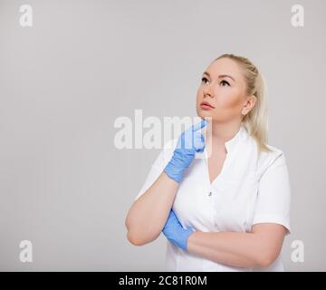 portrait de belle blonde femme médecin, infirmière ou cosmetologiste en uniforme blanc pensant à quelque chose sur fond gris avec l'espace de copie Banque D'Images