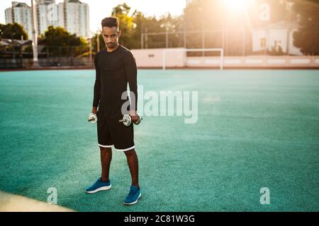 Jeune sportif afro-américain sérieux avec haltères regardant avec confiance l'appareil photo dans le stade Banque D'Images