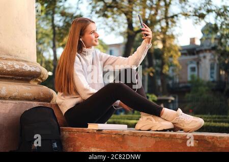 Jeune fille étudiante attrayante en chandail rêveur prenant selfie sur le téléphone portable pendant la pause d'étude en plein air Banque D'Images