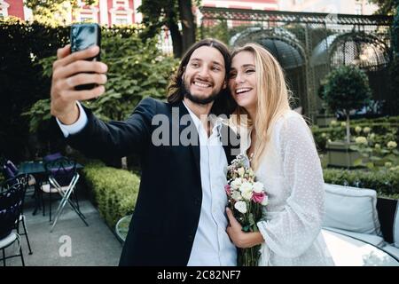 Jeune groom gai et mariée heureux de prendre selfie sur le téléphone portable dans le beau jardin du restaurant Banque D'Images