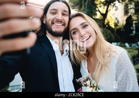 Jeune groom souriant et jeune mariée souriant prenant le selfie à l'extérieur Banque D'Images